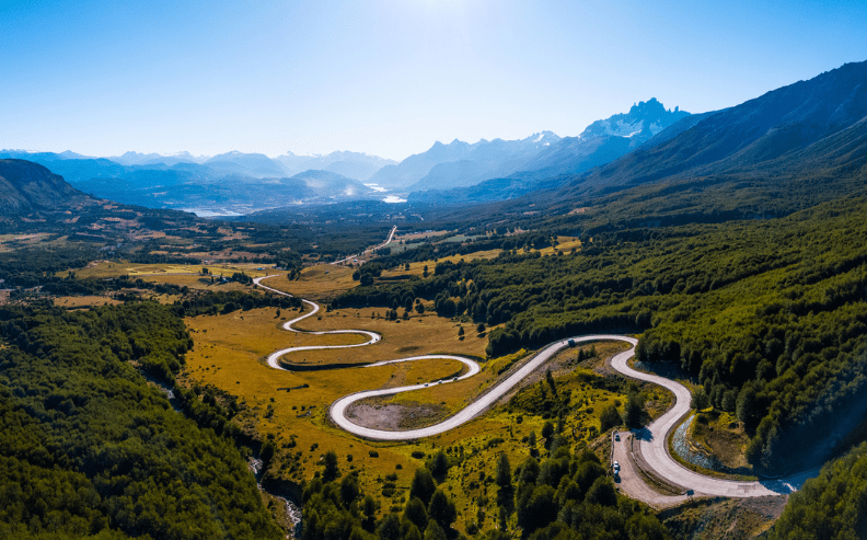 A winding mountain road curves through a lush green valley, surrounded by dense forests and distant snow-capped peaks under a clear blue sky. A car drives along the road, highlighting the scenic, remote landscape.