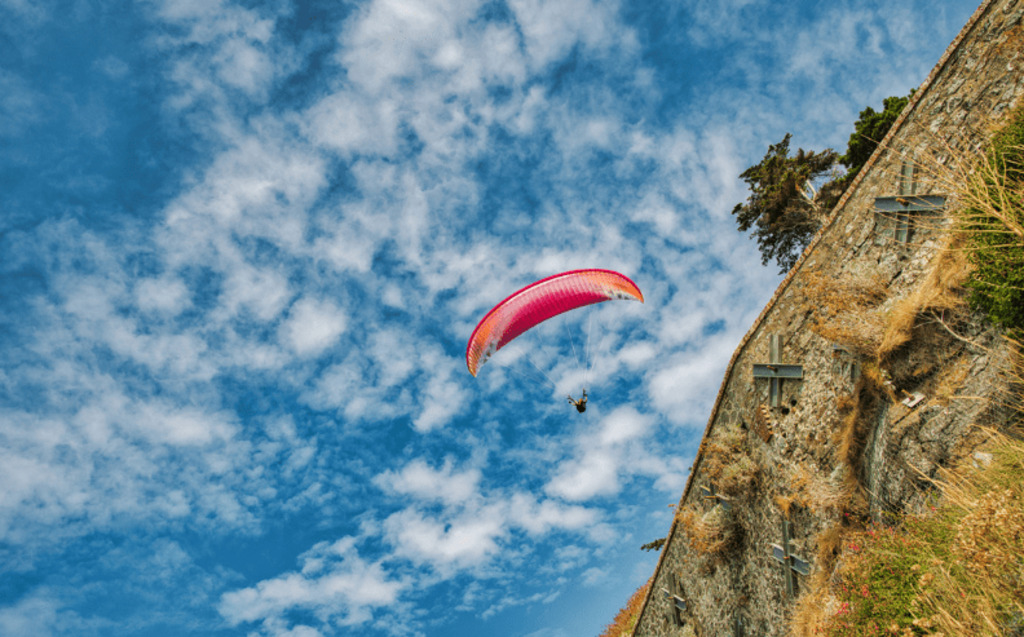 A person paraglides with a bright pink and orange parachute against a blue sky, soaring near a rocky cliff dotted with dry grass—experience the thrill and Request a Demo to See Radiant Logic In Action.