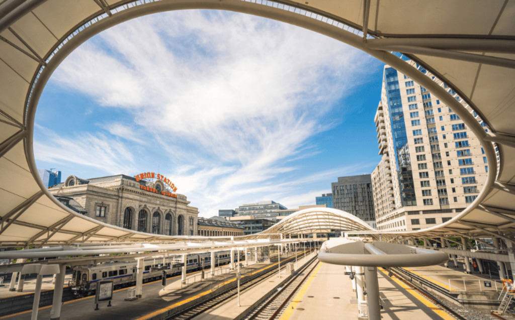 A wide view of train tracks and platforms at a modern outdoor station with curved white canopy structures; tall buildings and a historic station building are visible under a blue sky—Request a Demo to See Radiant Logic In Action.