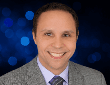 A man wearing a gray suit, lavender shirt, and striped tie is smiling at the camera. The background features blue bokeh lights.