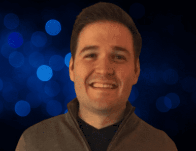 A man with short brown hair smiles at the camera, wearing a gray zip-up jacket. The background is dark with blue bokeh lights.