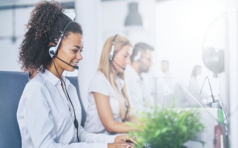 Three customer service representatives wearing headsets are sitting in a bright office, working at computers. The focus is on a smiling woman in the foreground. A green plant is visible on the desk.