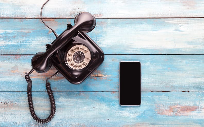 A black rotary dial telephone and a modern smartphone sit side by side on a light blue wooden surface, contrasting old and new communication devices.