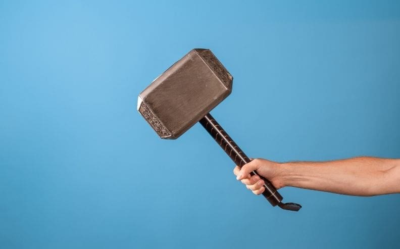 A person’s hand holds a large, gray, rectangular hammer with a short, patterned handle against a plain blue background.