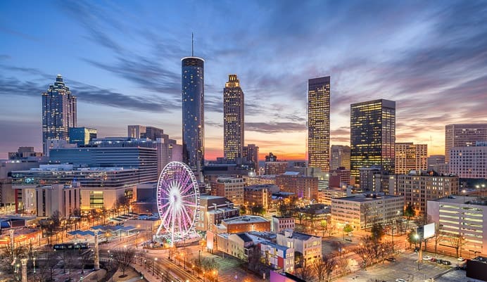 A view of downtown Atlanta at sunset, featuring illuminated skyscrapers, a lit Ferris wheel, and colorful clouds in the sky above the city skyline.