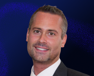 A man with short, neatly styled hair and light stubble, wearing a dark suit and white shirt, smiles in front of a dark blue background with a pattern of lighter blue dots.