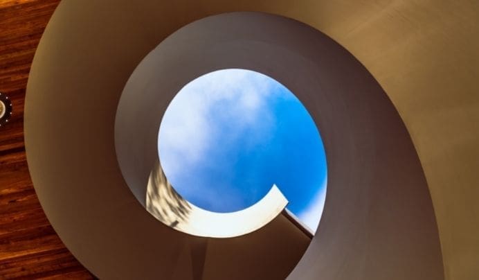 A spiral staircase, viewed from below, frames a circular patch of blue sky with light clouds at its center. The staircase structure is smooth and curves gracefully, with warm wooden flooring visible on the left side.