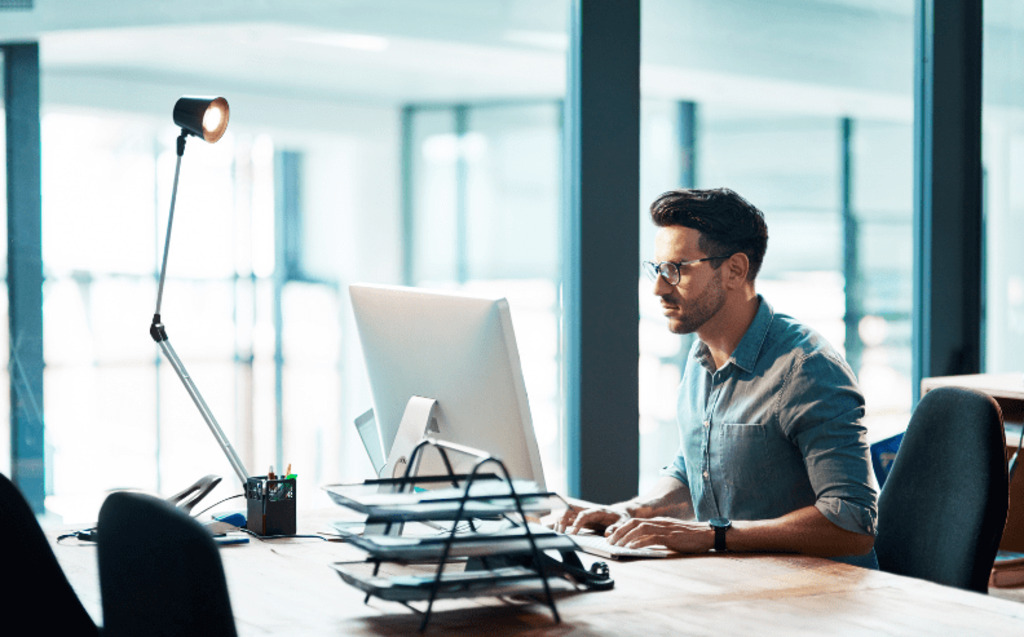 A man wearing glasses works at a computer in a modern, well-lit office, with documents and a desk lamp nearby. Large windows provide natural light and a cityscape view in the background.