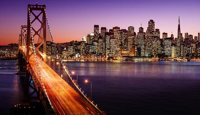 The Bay Bridge stretches into downtown San Francisco at dusk, with city skyscrapers illuminated and reflecting on the water beneath a purple and orange sky.