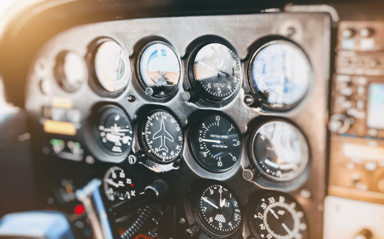 Close-up of a small aircraft cockpit showing various flight instruments and gauges, including an altimeter, airspeed indicator, vertical speed indicator, and attitude indicator, with sunlight shining in from the left.