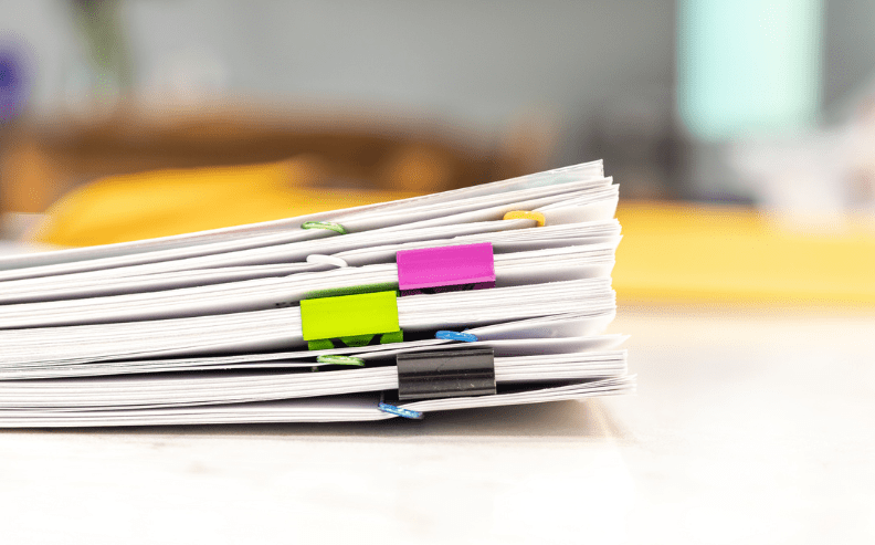 A stack of papers clipped together with colorful binder clips—green, pink, blue, and black—sits on a bright desk, with a blurred background.