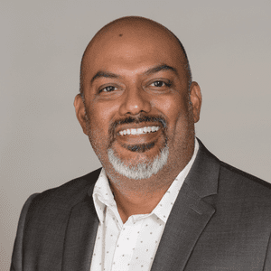 A smiling man with a shaved head and a salt-and-pepper beard, wearing a grey suit jacket over a white patterned shirt, posing in front of a plain light background.
