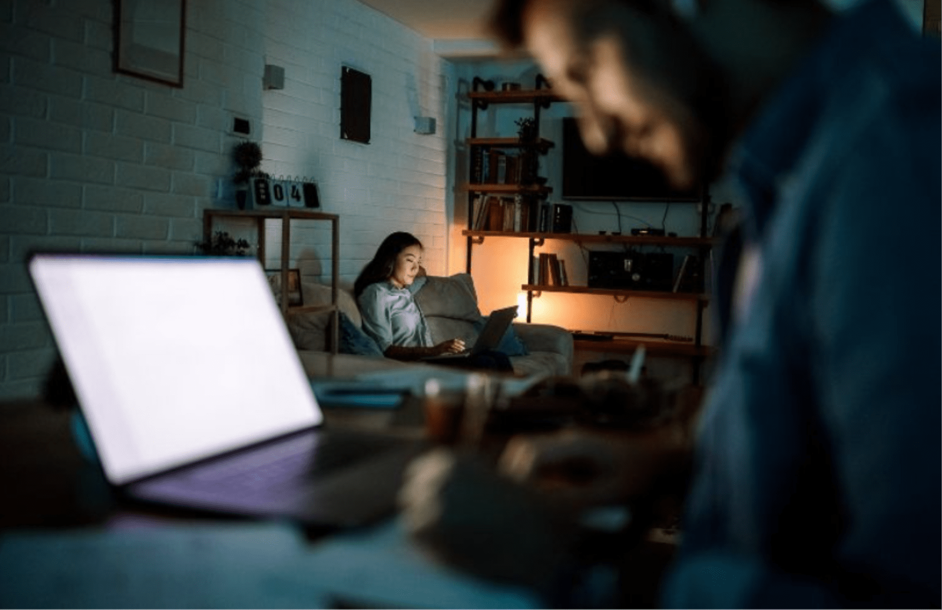 A woman sits on a couch using a laptop in a dimly lit room, while a blurred man works at a desk with another laptop in the foreground. The scene appears calm and focused.