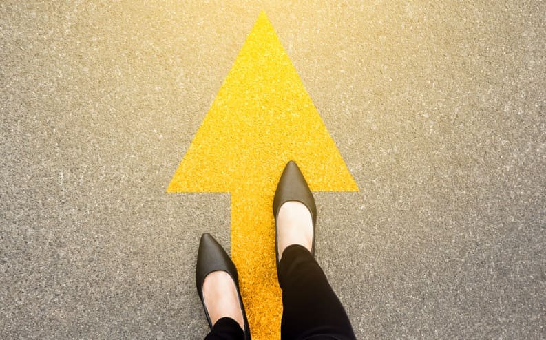 A person wearing black pants and black shoes stands on a paved surface with a large yellow arrow pointing forward, symbolizing direction or progress.