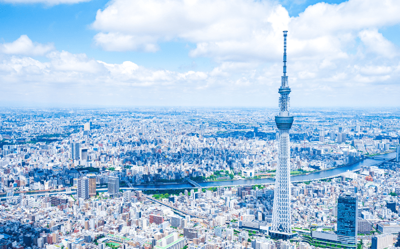 A panoramic view of Tokyo with the Tokyo Skytree tower rising prominently, surrounded by dense city buildings, a river, and a bright blue sky scattered with clouds.