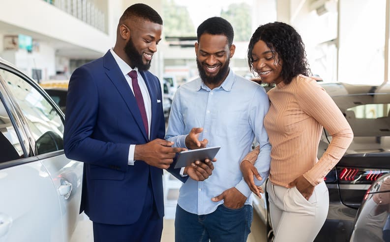 A car salesperson in a blue suit shows a tablet to a smiling man and woman standing beside a car in a bright showroom.