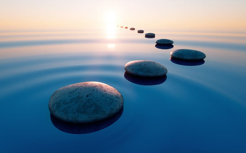 Smooth stones form a path across calm water, leading toward the horizon at sunrise or sunset. The sky is clear, and gentle ripples surround each stone, creating a peaceful and serene atmosphere.