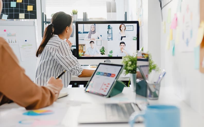 Two people sit at a desk in a modern office, participating in a video conference with six colleagues displayed on a computer monitor. Laptops, charts, and stationery are visible on the desk.
