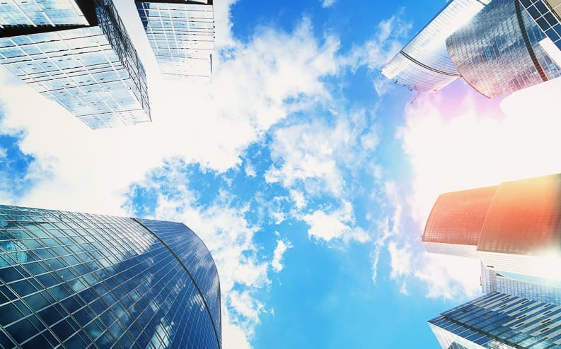 View looking up at modern glass skyscrapers reflecting sunlight, with a bright blue sky and scattered clouds in the background.