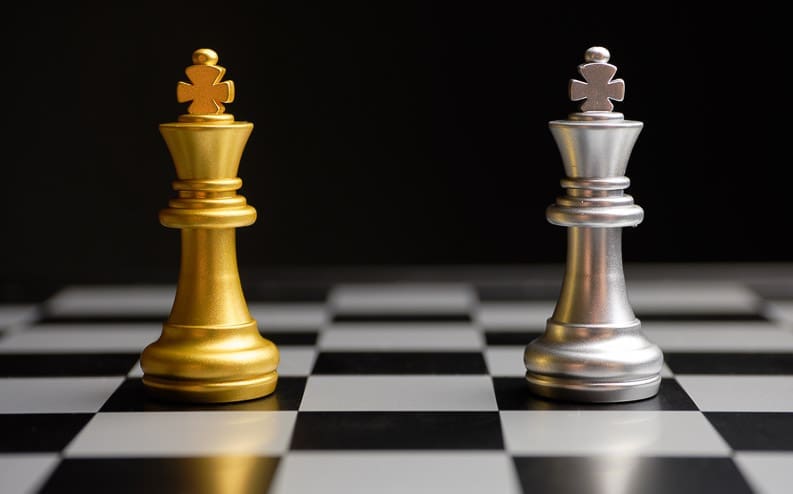 Two chess kings, one gold and one silver, stand on a black and white chessboard against a dark background.