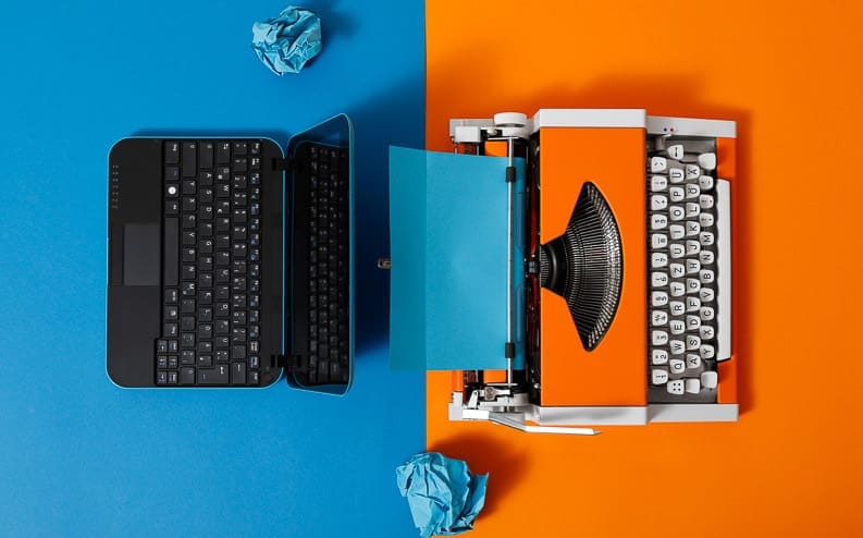 A laptop and a typewriter are placed side by side on a split blue and orange background, with crumpled paper balls nearby, symbolizing the contrast between old and modern writing technology.