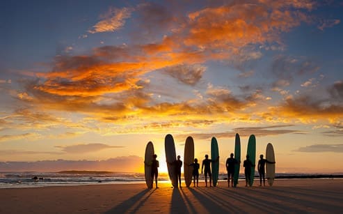 Seven people stand in a row on a beach at sunset, each holding a surfboard upright. The sky is filled with dramatic orange and yellow clouds above the calm ocean.