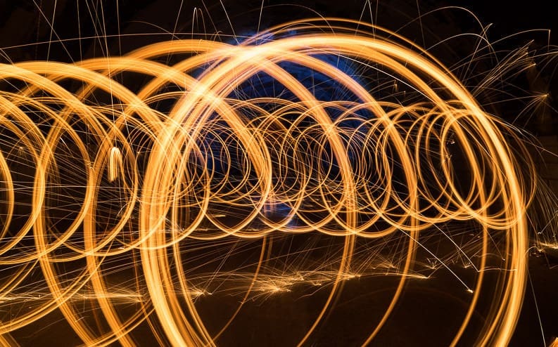 Long-exposure photo of swirling orange and yellow light trails forming overlapping circular patterns against a dark background, creating a dynamic, abstract effect.