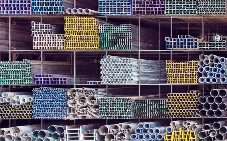 Stacks of colorful plastic and metal pipes of various shapes and sizes are neatly arranged in a large shelving unit, creating a geometric and organized pattern. The pipes are grouped by color and type.