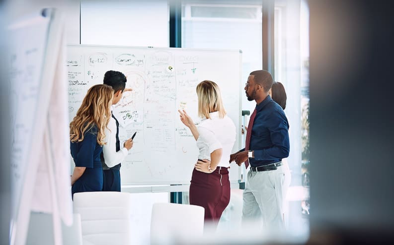 A group of five people stand around a whiteboard covered with notes and diagrams, discussing and brainstorming ideas in a modern office setting.