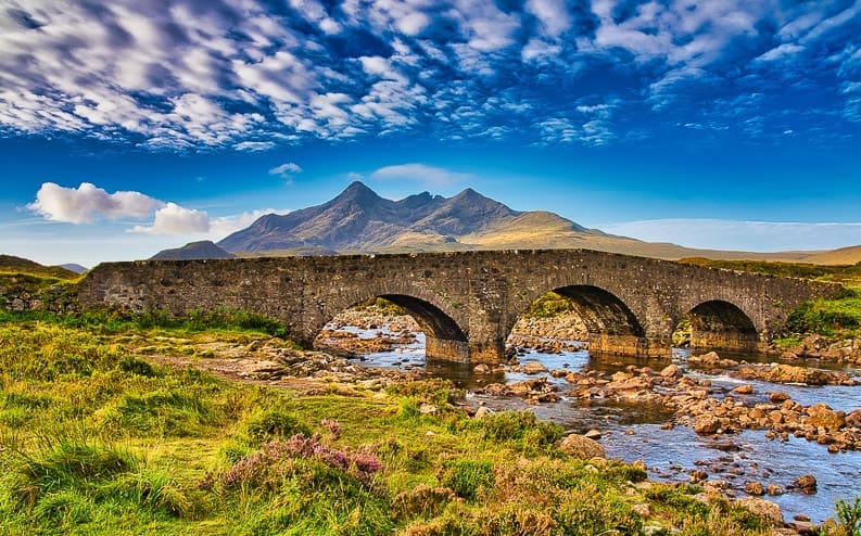A historic stone bridge with three arches spans a rocky river, set against a backdrop of green hills and jagged mountains under a vibrant blue sky with scattered clouds.