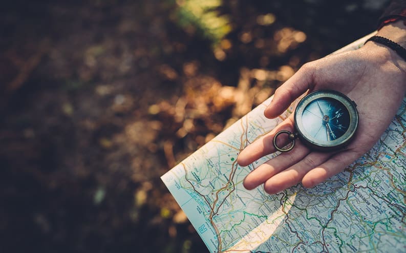 A person holds a vintage compass in their hand over a detailed map, surrounded by blurred outdoor foliage and sunlight.