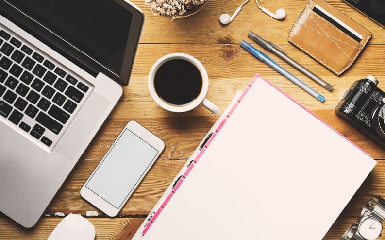 A wooden desk with a laptop, smartphone, blank notebook, coffee cup, pens, camera, wallet, earphones, and bowl of popcorn, arranged in a flat lay style.