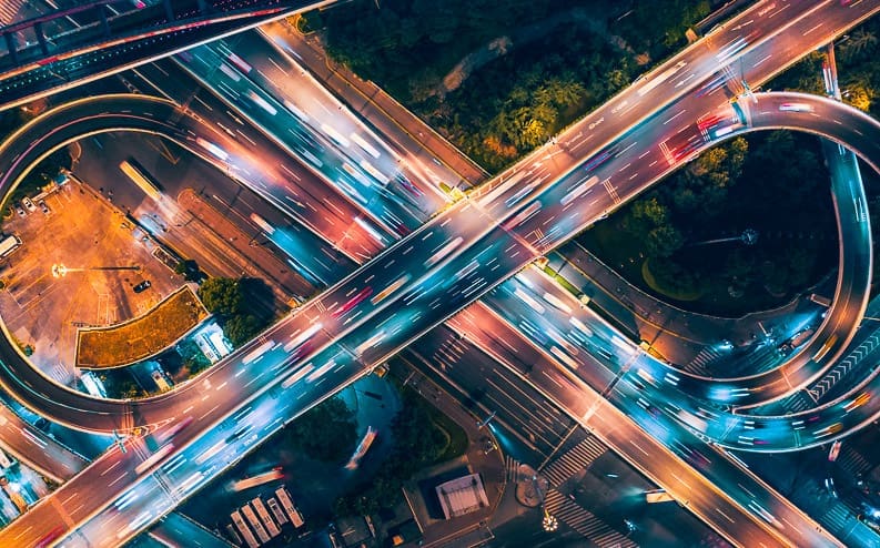 Aerial view of a busy multi-layered highway interchange at night, with blurred car lights creating colorful streaks and intersecting roads illuminated by streetlights.
