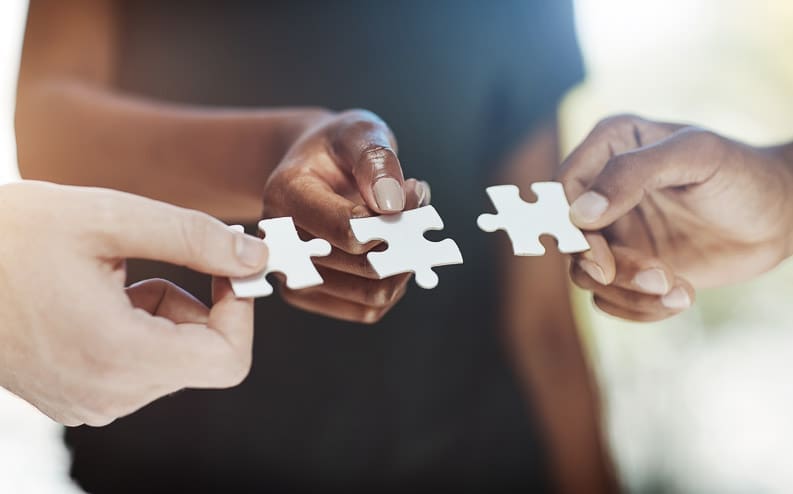 Three people hold white puzzle pieces close together, their hands meeting in the center of the image, symbolizing teamwork and collaboration.