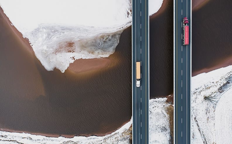 Aerial view of two parallel highways over a partially frozen river, with one truck on each road, surrounded by snow-covered ground.