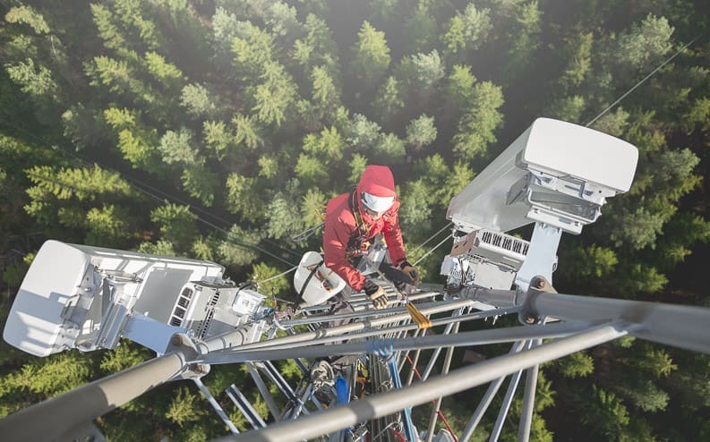 A worker wearing safety gear climbs a tall communications tower high above a forest, surrounded by large antennas and equipment, with trees visible far below.