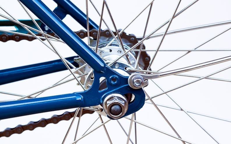 Close-up of the rear wheel of a bicycle, showing metal spokes, blue frame, chain, and axle nut against a light background.