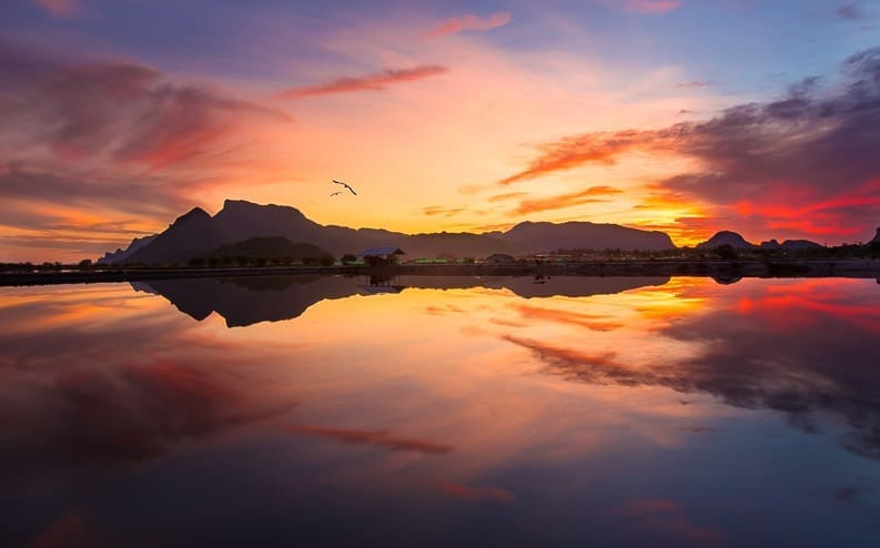 A vibrant sunset sky with orange, pink, and purple hues is reflected in calm water, with silhouetted mountains and a flying bird completing the serene landscape.