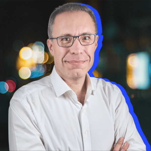 A man wearing glasses and a white shirt stands with his arms crossed, smiling at the camera. The background is blurred with colorful lights, and there is a blue outline around his figure.