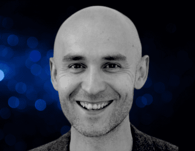 A smiling bald man in a dark jacket is shown against a dark blue background with blurred light spots.