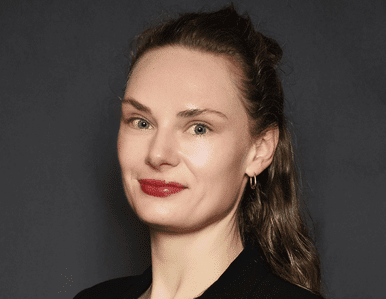 A woman with light skin, long wavy brown hair, and red lipstick, wearing a black top and small hoop earrings, poses against a dark background, looking at the camera with a slight smile.