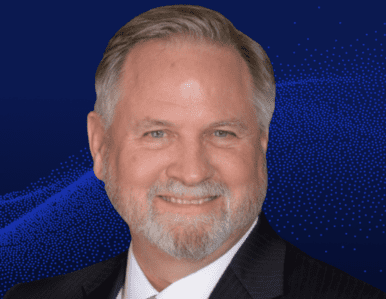 A middle-aged man with gray hair and a beard, smiling and wearing a dark suit and white shirt, in front of a dark blue background with light blue dotted patterns.