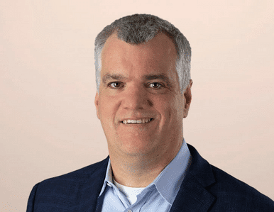 A middle-aged man with short gray hair is smiling. He is wearing a dark blue blazer, a light blue shirt, and a white undershirt, posed against a plain, light-colored background.