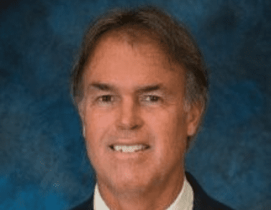 A middle-aged man with brown hair, wearing a dark suit and white shirt, smiling in front of a blue textured background.