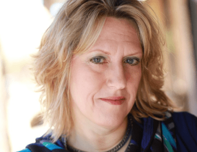 A woman with wavy blonde hair and light makeup looks at the camera with a slight smile. She is wearing a blue and green top and a necklace, and the background is softly blurred.