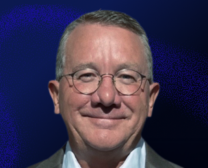 A middle-aged man with short gray hair, glasses, and a slight smile, wearing a dark suit jacket and white shirt, is pictured against a dark blue background.