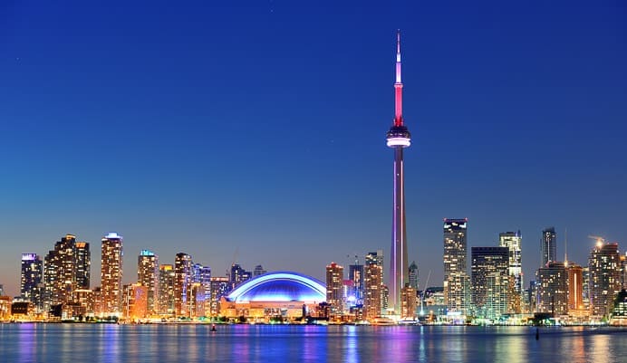 Toronto skyline at dusk, featuring illuminated skyscrapers, the CN Tower with red and white lights, and the Rogers Centre glowing blue, all reflected in the calm water below.