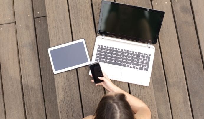 A person sits on a wooden floor holding a smartphone, with a tablet and an open laptop placed in front of them. The screens of all devices are turned off or blank.