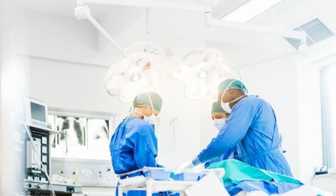Two surgeons in blue scrubs and masks operate on a patient in a brightly lit, sterile operating room with surgical lights overhead and medical equipment nearby.