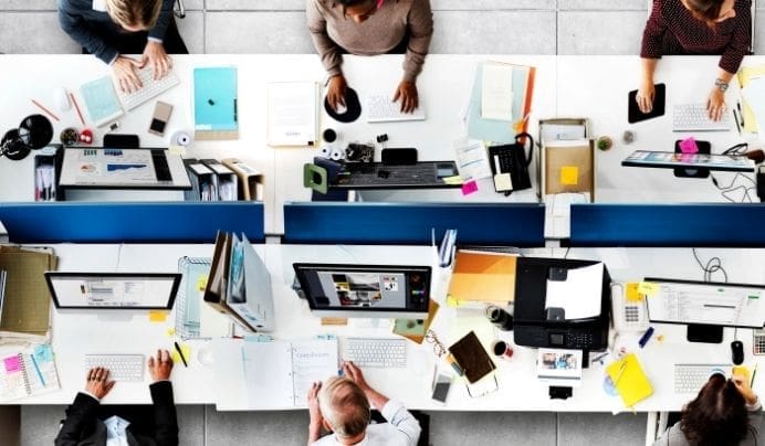 Overhead view of six people working at desks with computers, documents, coffee cups, and office supplies, arranged in two rows, creating a busy office environment.